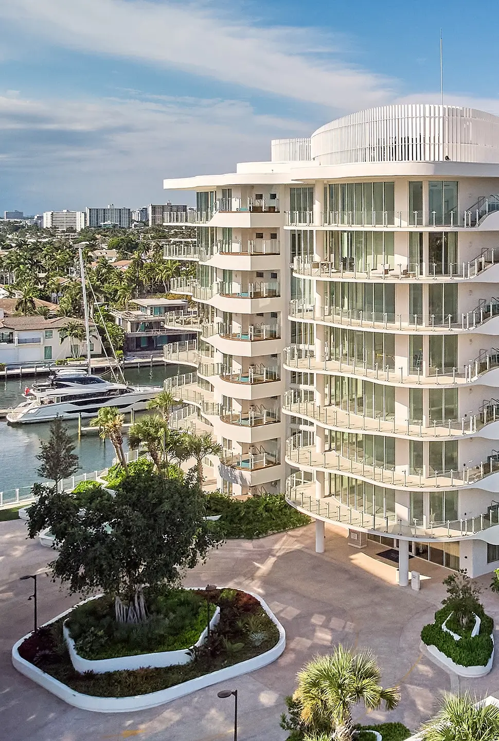 White curved hotel by a marina with glass balconies and palm trees amid boats and calm water.
