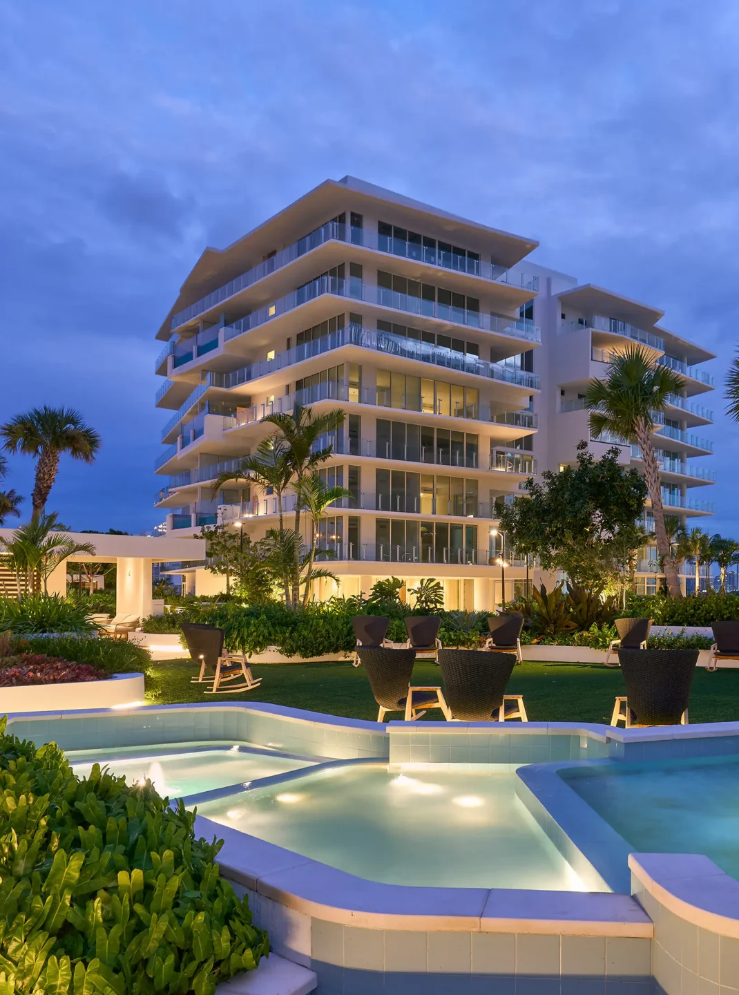 Modern glass-fronted apartment/hotel building with stacked balconies, poolside lounge area, and palm trees at dusk.