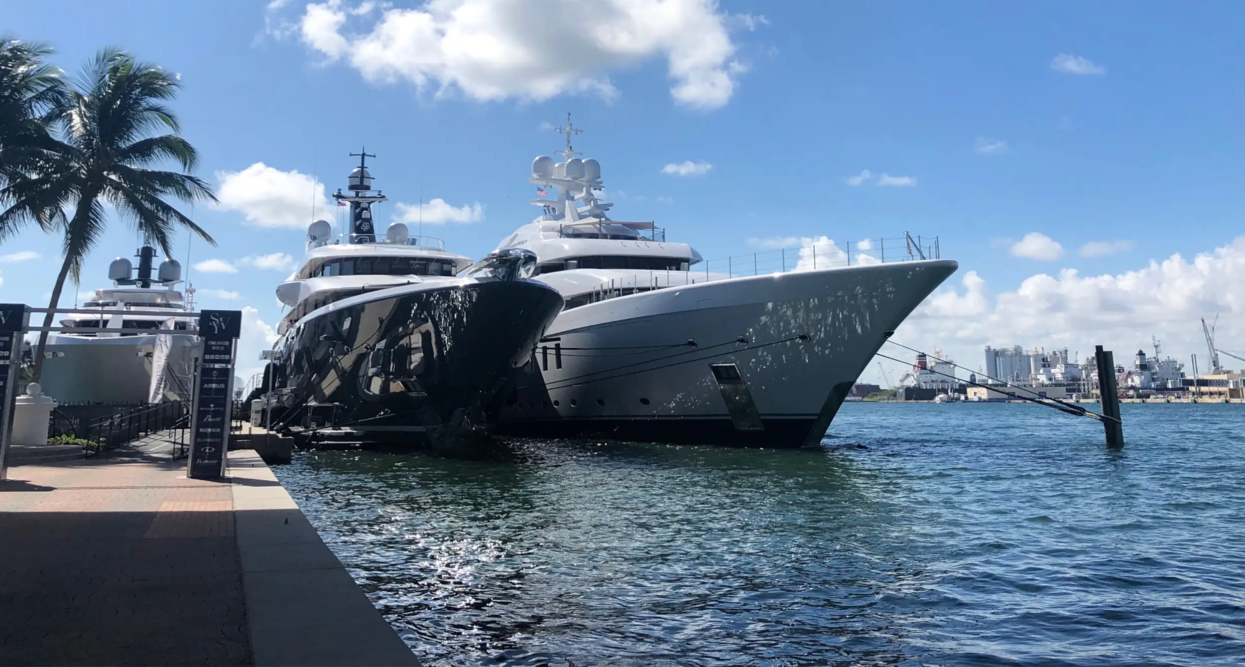 Large white luxury yacht docked at a sunny marina with palm trees nearby and a blue sky above.